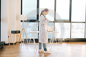 Portrait of thoughtful female doctor in white coat standing on background of window in sunny day in light medical clinic