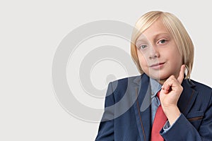 Portrait of thoughtful little boy with blond hair in blue jacket and red tie, keeps finger on chin, ponders something, isolated