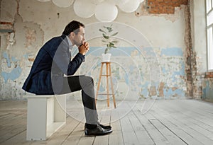 Portrait of thoughtful man, businessman sitting in empty big room and thinking about new company opening. Professional