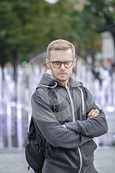 Portrait of a thoughtful man in glasses 25-30 years old with a backpack on his shoulder against a blurry background of city founta
