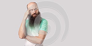 Portrait of thoughtful middle aged bald man with long beard in light green t-shirt standing and looking away, thinking about