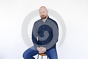 Portrait of thoughtful middle-aged man in blue shirt, jeans sitting on stool, interlacing fingers on white background.