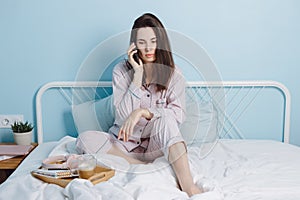 Portrait of thoughtful serious confident brunette friendly smart girl sitting on bed talking on phone, drinking coffe