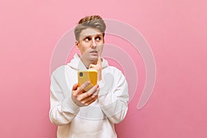 Portrait of thoughtful student boy in white hoodie on pink background holding smartphone, looking away at copy space with pensive