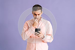 Portrait of thoughtful young man holding phone with doubtful and skeptical expression on pink isolated background.