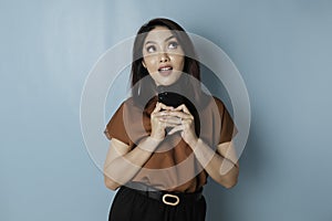 Portrait of a thoughtful young woman wearing brown shirt looking aside while holding smartphone  over blue background