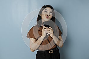 Portrait of a thoughtful young woman wearing brown shirt looking aside while holding smartphone  over blue background
