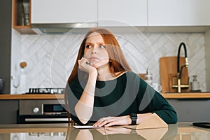 Portrait of thoughtful young woman writing handwritten letter sitting at table with envelope in kitchen room with modern