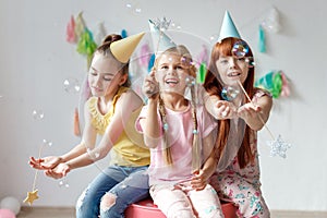 Portrait of three beautiful girls wear festive caps, play with bubbles, sit together on chair, celebrate birthday, being