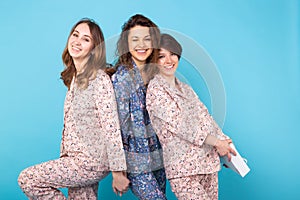 Portrait of three beautiful young girls wearing colorful pyjamas having fun during sleepover isolated over blue
