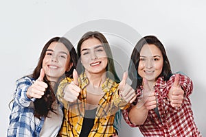 Portrait of three beautiful young happy females smiling joyfully showing thumbs up on white