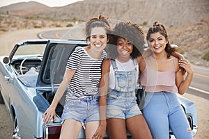 Portrait Of Three Female Friends Sitting In Trunk Of Classic Car On Road Trip