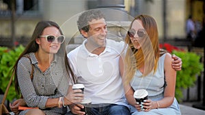 Portrait of three friends drinking coffee sitting on the bench outdoors in the city during warm summer day