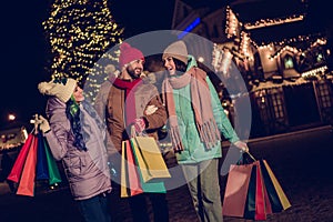 Portrait of three peaceful positive buddies hold shop bags walking speak communicate evergreen garland lights outdoors