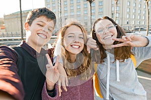 Portrait of three teen friends boy and two girls smiling and taking a selfie outdoors. City background, golden hour