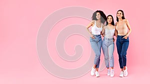 Portrait of three young diverse ladies friends embracing and posing over pink background, panorama with free space