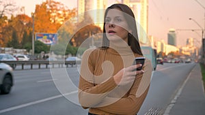 Portrait of tired brunette caucasian girl nervously standing near road with phone in hands.