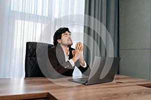 Portrait of tired businessman in suit sitting at office desk with laptop and pray to God ask for good luck in business