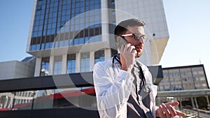 Portrait of tired exhausted doctor having phone call outside hospital in morning. COVID-19, Coronavirus pandemic