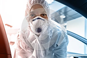 Portrait of tired exhausted female doctor, scientist or nurse wearing face mask and biological hazmat protective suit open car