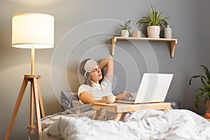Portrait of tired exhausted woman wearing sleep mask sitting with laptop in bed at home and working online, feeling pain in neck,