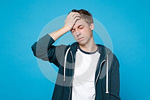 Portrait of tired exhausted young man in casual clothes keeping eyes closed, putting hand on head isolated on blue