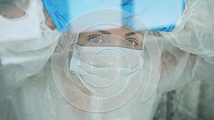 Portrait tired isolated medic woman with green eyes in white protective suit, medical face mask, blue gloves leaning head