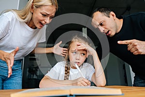 Portrait of tired little girl doing homework sitting at desk at home on background of angry parents scolding together