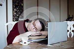Portrait of tired middle-aged woman sitting at table near laptop, sleeping on crossed arms on pile of book sat home.