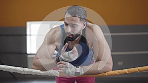 Portrait of tired Middle Eastern boxer resting leaning on ropes. Confident strong man having break in training on boxing