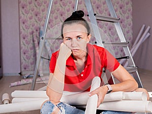 Portrait of a tired and sad woman sitting on the floor with rolls of Wallpaper in her hands.