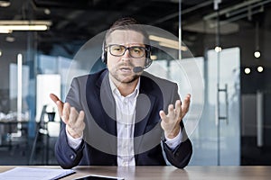 Portrait of a tired and upset young man in a headset sitting in the office in front of the camera and unhappy talking on