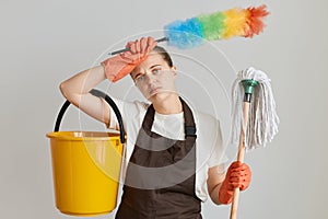 Portrait of tired woman wearing brown apron and white t shirt cleaning her flat, holding mop, ppduster and bucket, keeping hand on