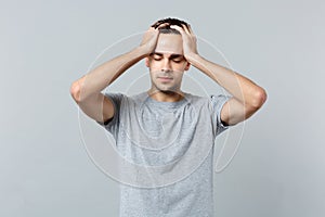 Portrait of tired young man in casual clothes keeping eyes closed, putting hands on head isolated on grey wall