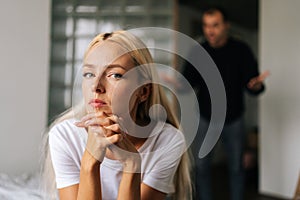 Portrait of tired young woman sadness looking at camera sitting on bed on background of aggressive husband shouting on