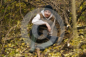 Portrait of traveler hiker man sitting on spread out raincoat in thicket under tree in forest on overcast rainy day to