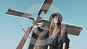 Portrait of traveller woman near old windmill. Incredible cinematic low angle. Girl in hat in front of farm mill. 4K