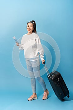 Portrait of trendy young girl standing with suitcase and holding passport with tickets, over blue background