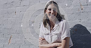 Portrait of a trendy young woman smiling and laughing with arms crossed against a wall on a sunny day. Happy