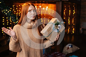 Portrait of troubled redhead young woman holding in hand beautiful gift box with Christmas present.