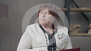 Portrait of troubled thoughtful Caucasian senior woman sitting with laptop at home thinking. Redhead female mature