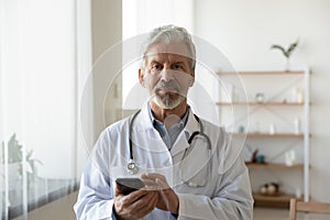 Portrait of trusted old mature doctor with cellphone in hands.