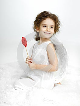 Portrait of turned little girl with angelic smiling in white dress and wings, holding red heart, over white background.