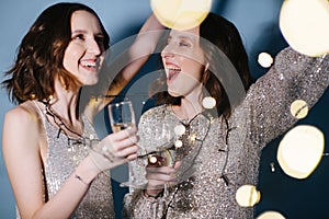 Twin sisters in shiny evening dresses celebrate with glasses of sparkling wine