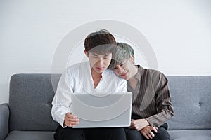 Portrait of two Asian young happy gay lovers sitting snuggle and using laptop computer together on sofa