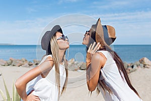 Portrait of two attractive brunette and blond girls with long hair are standing on the beach near sea. They wear hats