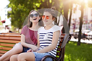 A portrait of two attractive young friends who have rest on wooden bench in local park after long walk. Laughing girls have lots