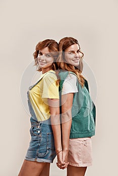 Portrait of two attractive young girls, twin sisters in casual wear smiling at camera, holding hands, posing together