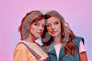 Portrait of two attractive young girls, twin sisters looking at camera, posing together isolated over pink background