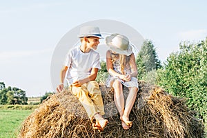 Portrait of two barefoot children boy and girl sitting on haystack in field. Smiling and laughing kids wearing hats.